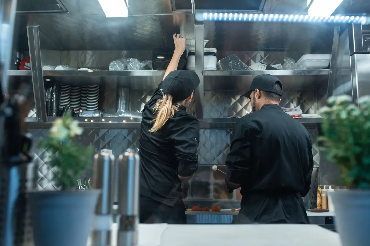 Two cooks in black chef jackets working a stainless steel line under a pass shelf