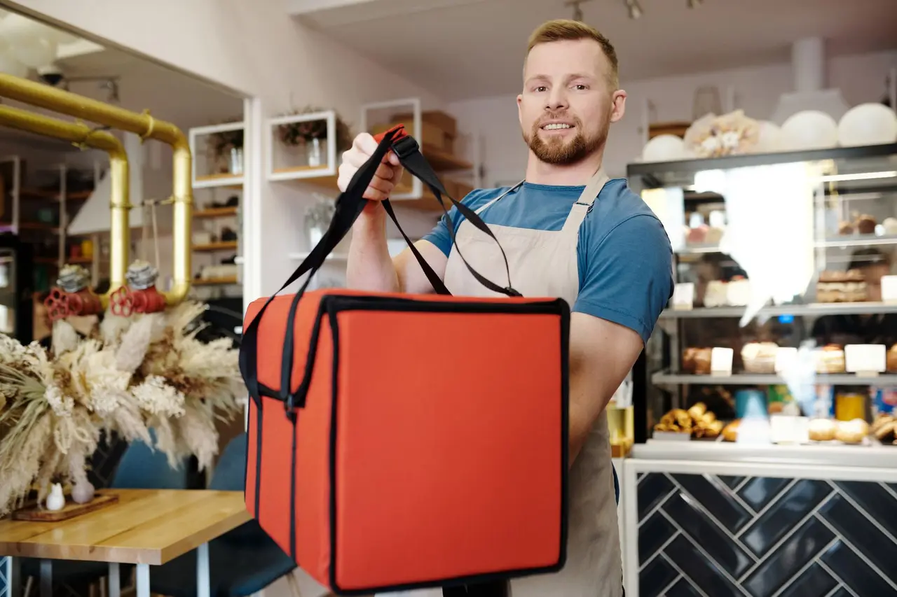 A kitchen worker in an apron handing over an insulated delivery bag