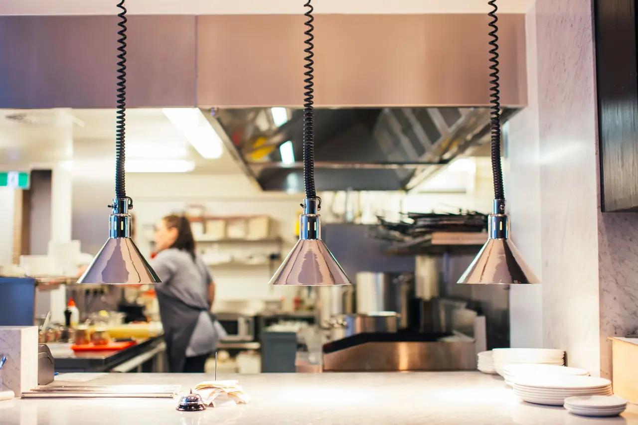 Three heat lamps over an empty kitchen pass with a ticket spike, a service bell and stacked plates, a cook working the line behind